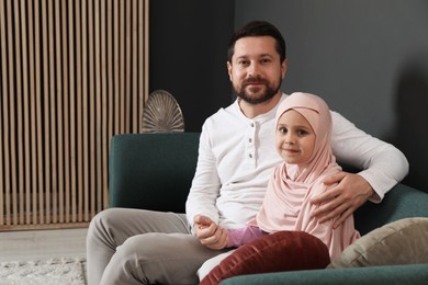 Photo of Muslim man and his daughter sitting on sofa at home, space for text