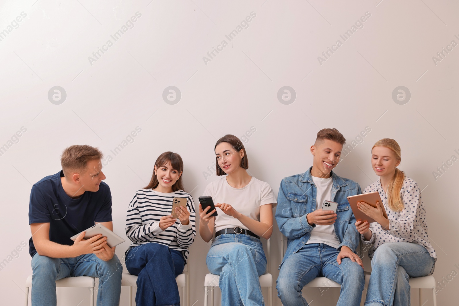 Photo of Group of people using different gadgets on chairs near white wall indoors. Modern technology