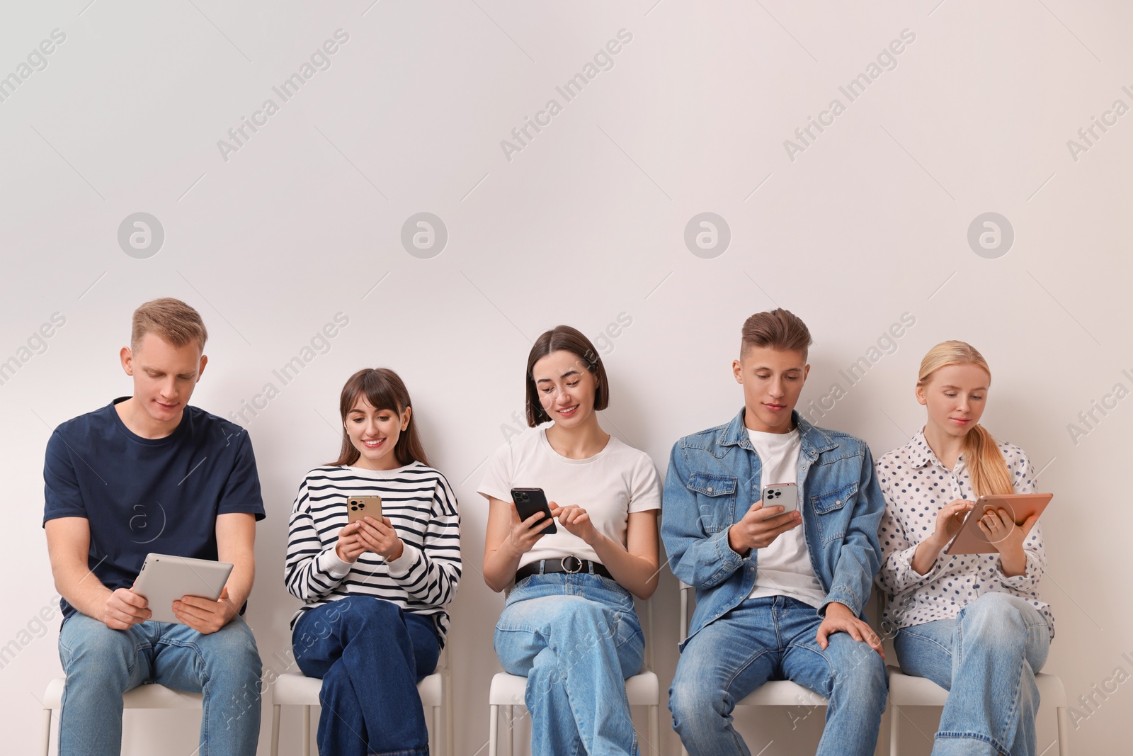 Photo of Group of people using different gadgets on chairs near white wall indoors. Modern technology