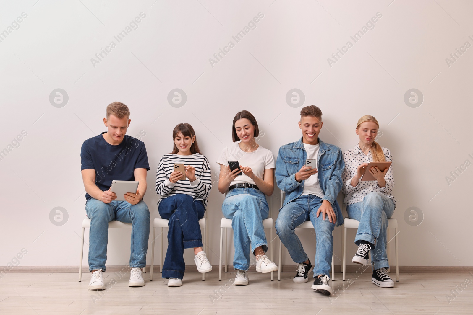 Photo of Group of people using different gadgets on chairs near white wall indoors. Modern technology