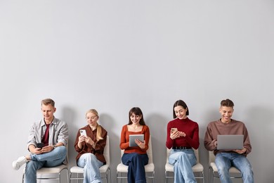 Photo of Group of people using different gadgets on chairs near light grey wall indoors. Modern technology