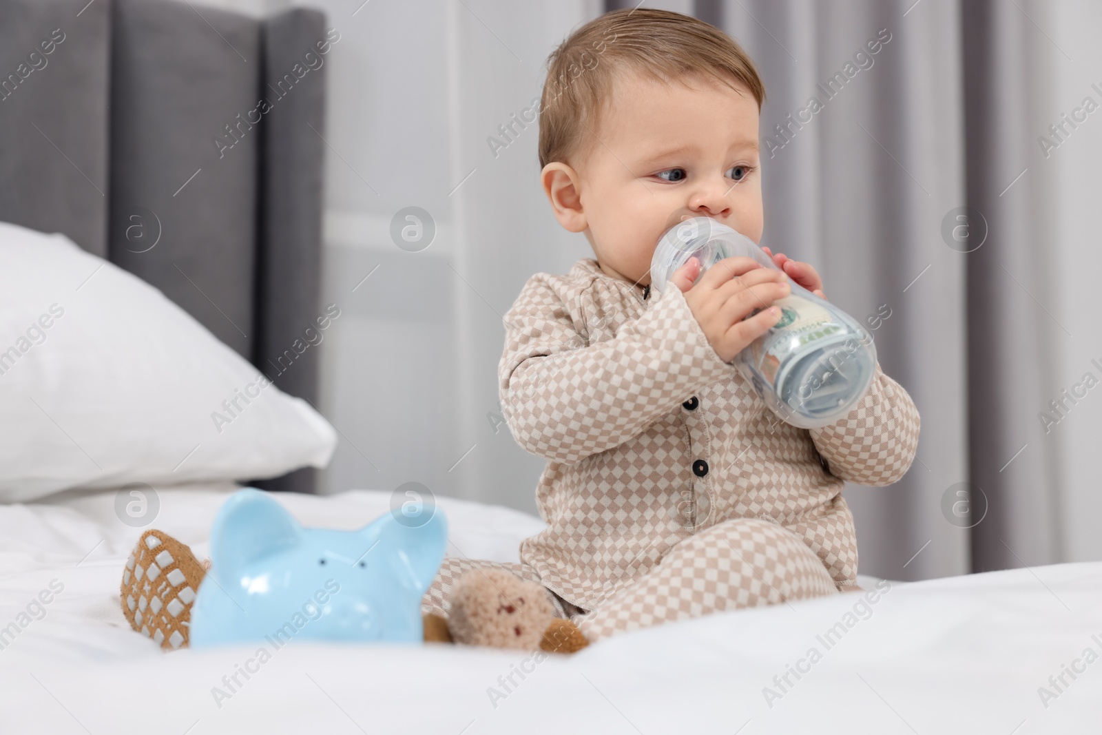 Photo of Cute little baby with money in bottle and piggybank on bed at home