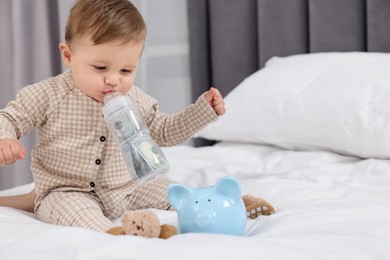 Photo of Cute little baby with money in bottle and piggybank on bed at home. Space for text