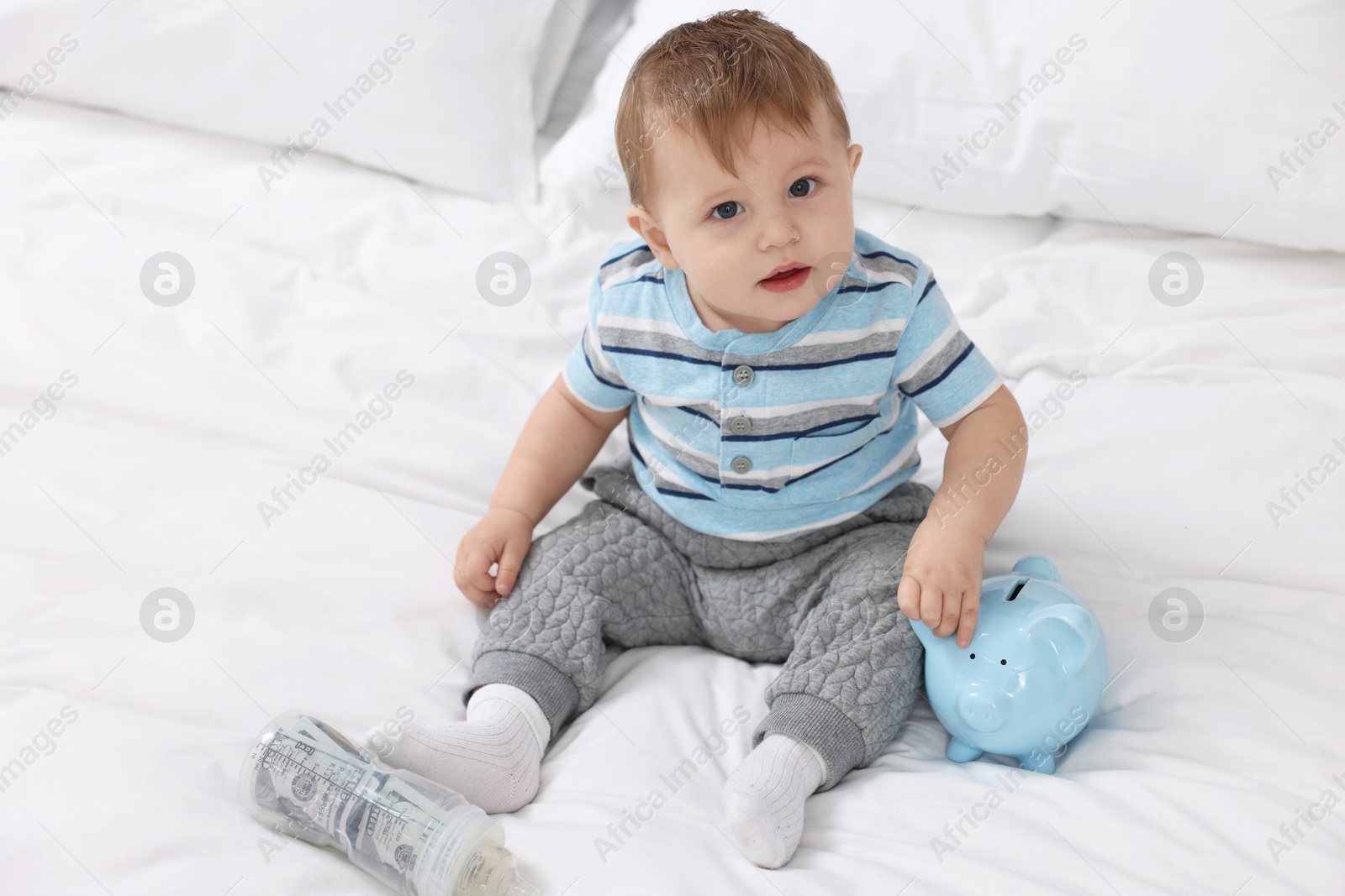 Photo of Cute little baby with money in bottle and piggybank on bed at home