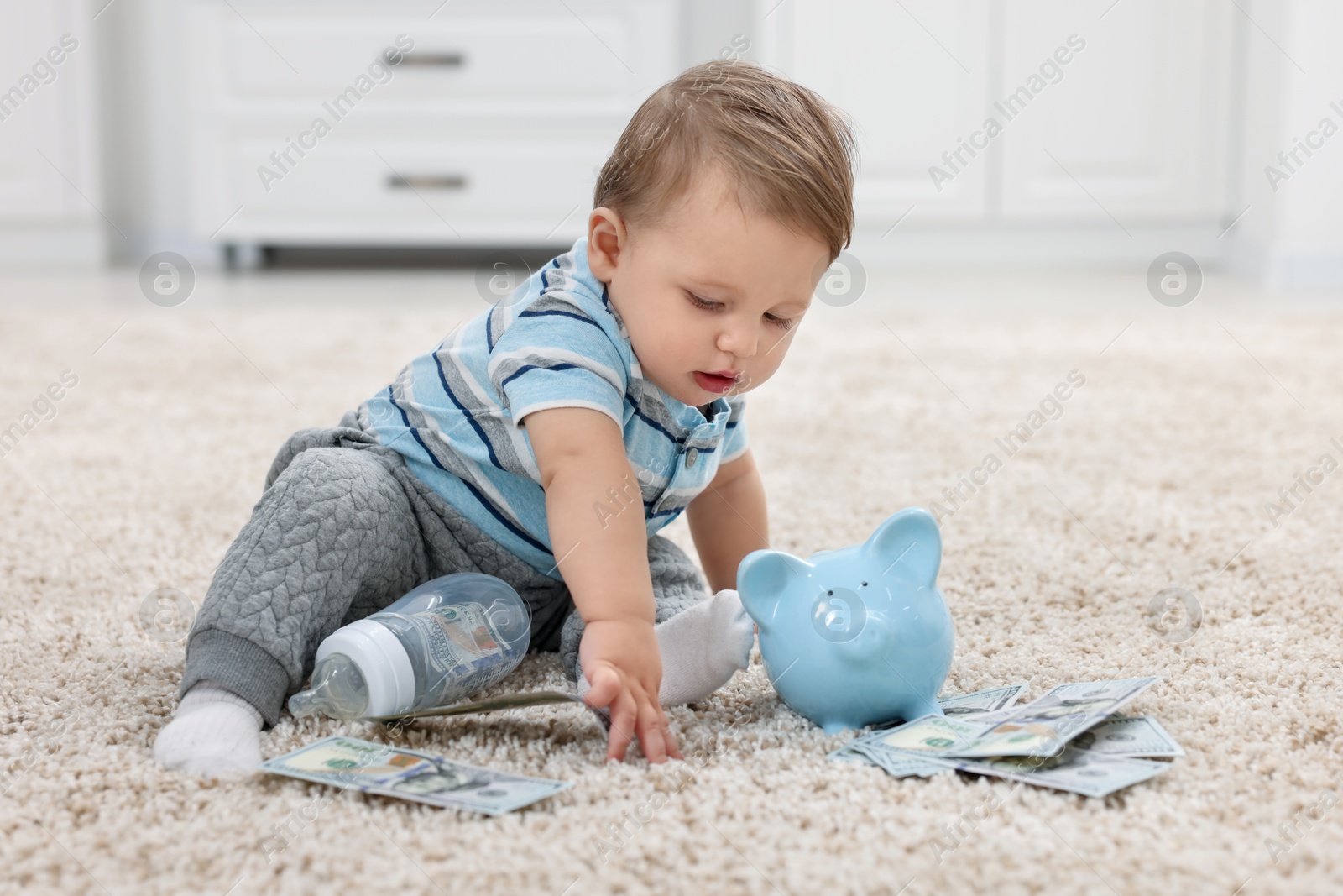 Photo of Little baby with bottle, money and piggybank on floor at home