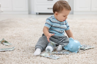 Photo of Little baby with bottle, money and piggybank on floor at home