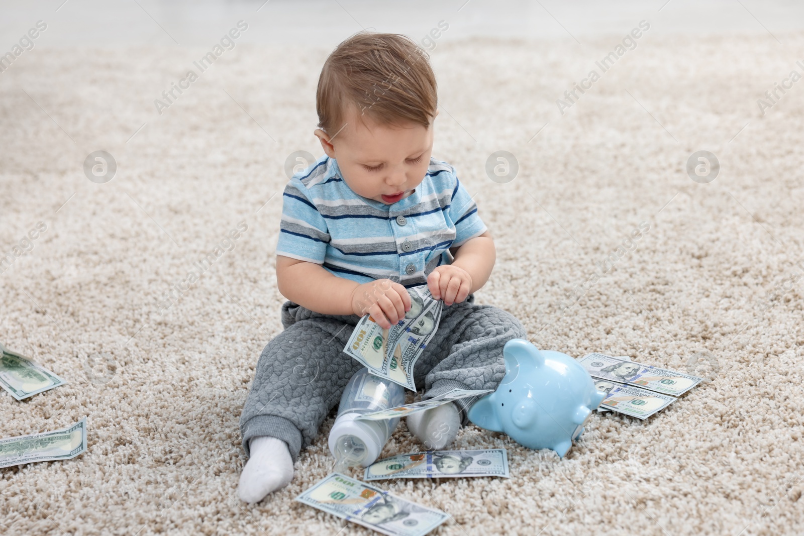 Photo of Little baby with bottle, money and piggybank on floor at home