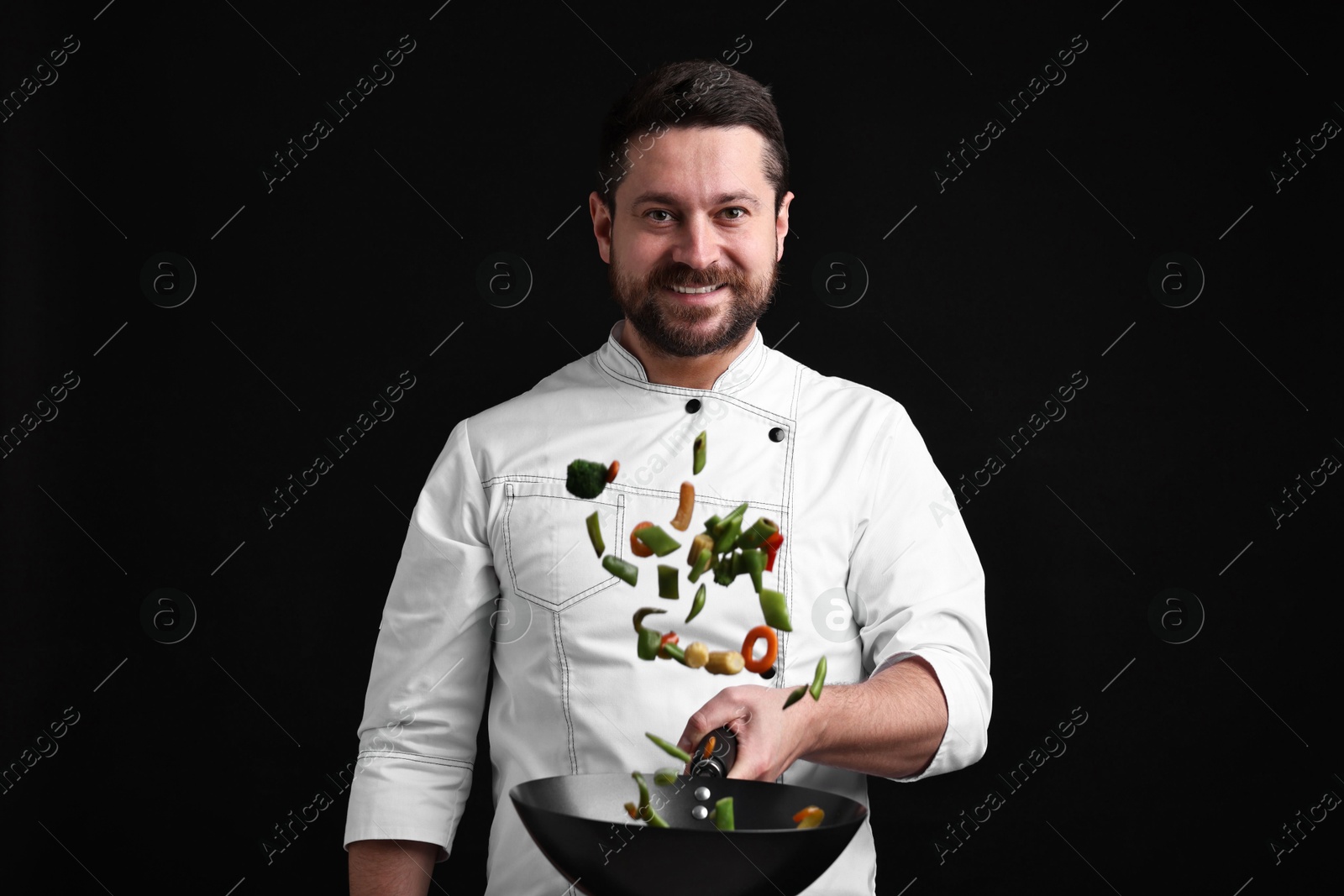 Photo of Professional chef mixing vegetables in wok on black background