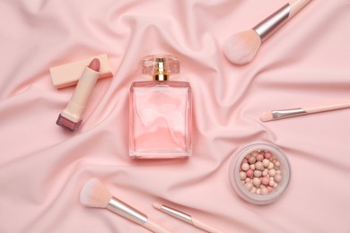 Photo of Bottle of perfume, ball powder, lipstick and makeup brushes on pink silk fabric, flat lay