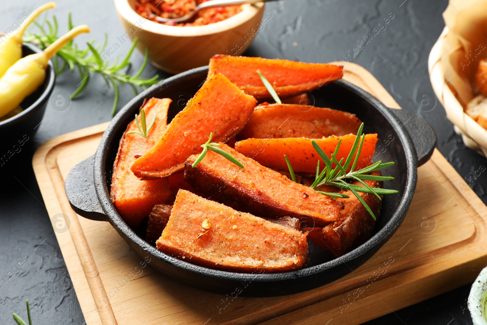 Photo of Pieces of tasty cooked sweet potato with rosemary in baking dish served on grey textured table, closeup