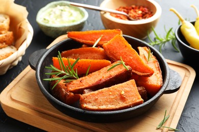 Photo of Pieces of tasty cooked sweet potato with rosemary in baking dish served on grey textured table, closeup