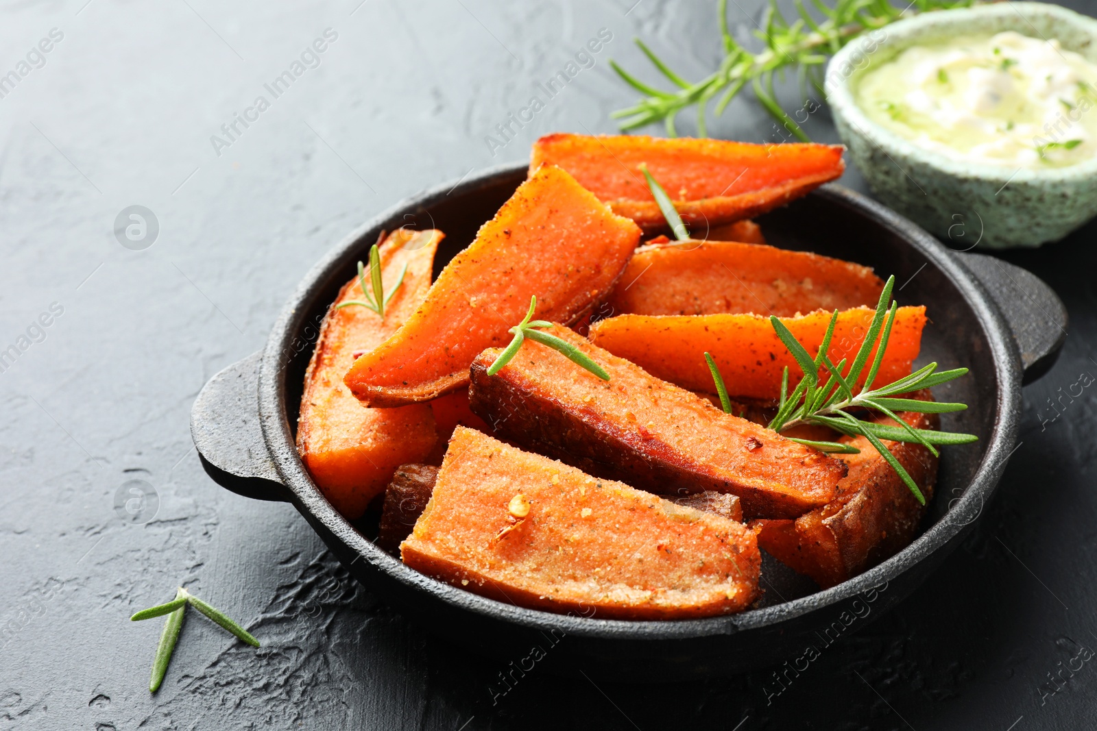 Photo of Pieces of tasty cooked sweet potato with rosemary in baking dish served on grey textured table, closeup