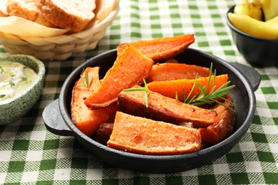 Pieces of tasty cooked sweet potato with rosemary in baking dish served on checkered tablecloth