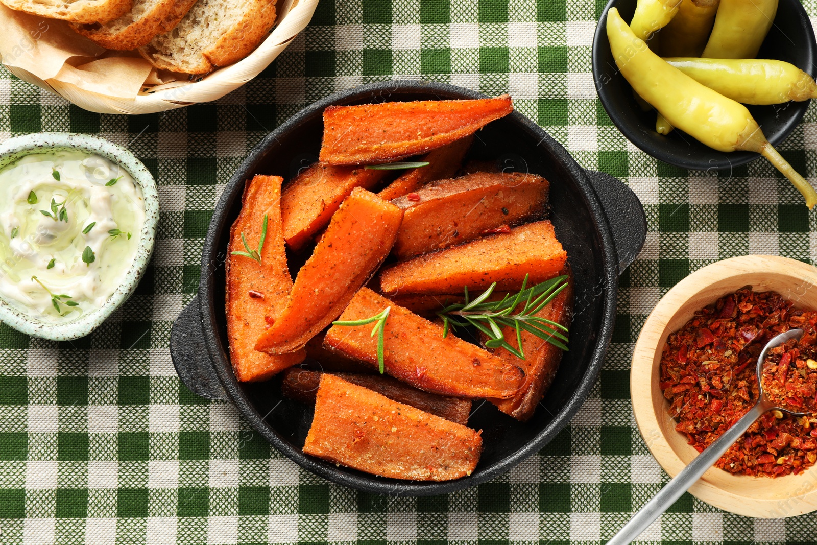 Photo of Pieces of tasty cooked sweet potato with rosemary in baking dish served on checkered tablecloth, flat lay