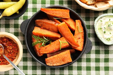 Pieces of tasty cooked sweet potato with rosemary in baking dish served on checkered tablecloth, flat lay