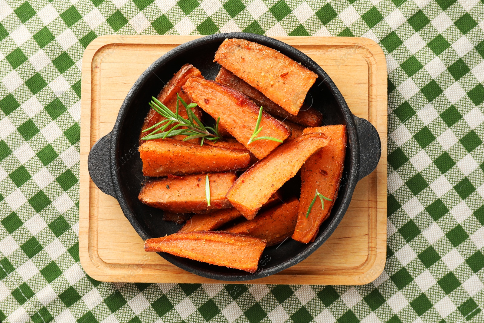 Photo of Pieces of tasty cooked sweet potato with rosemary in baking dish on checkered tablecloth, top view