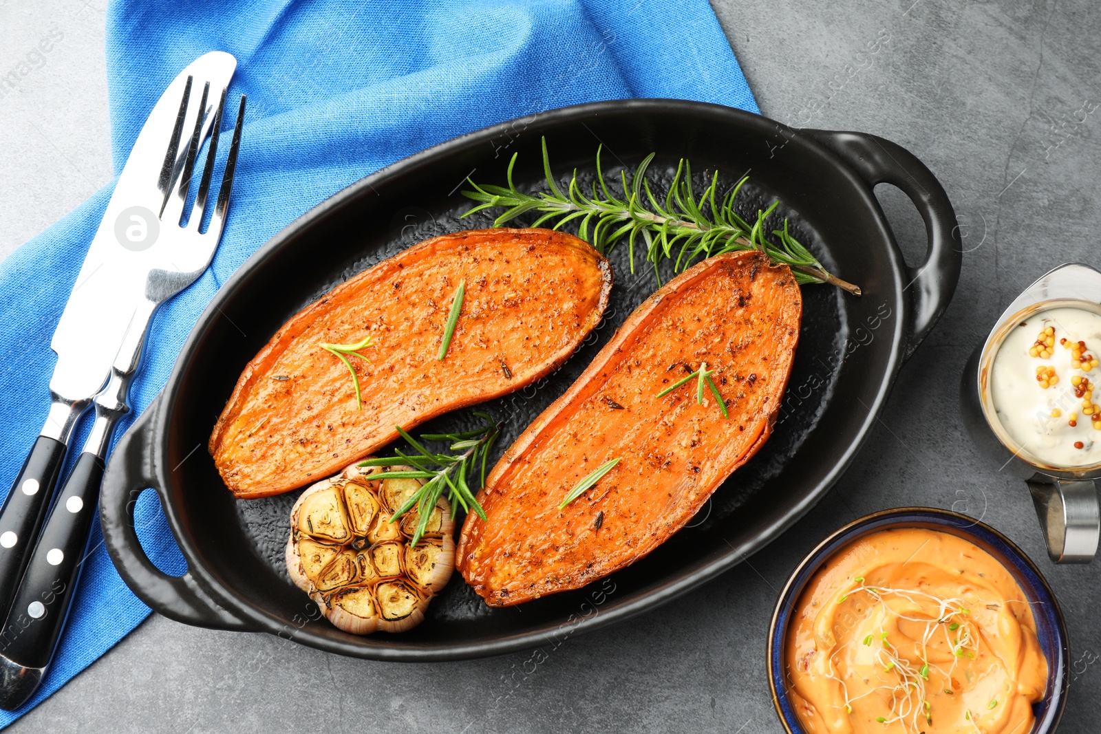 Photo of Halves of tasty cooked sweet potato with rosemary and garlic in baking dish served on grey textured table, flat lay