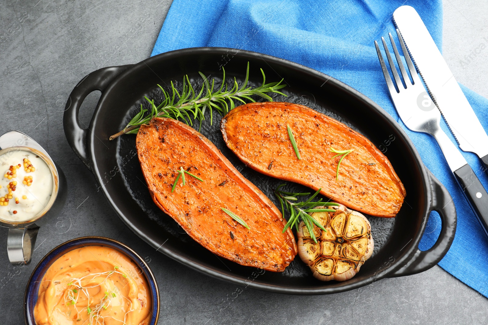 Photo of Halves of tasty cooked sweet potato with rosemary and garlic in baking dish served on grey textured table, flat lay