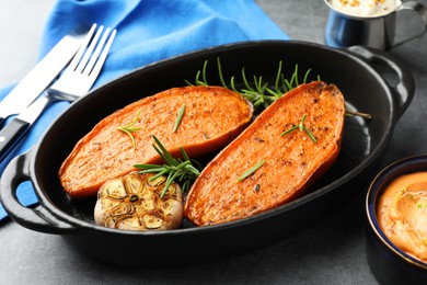 Photo of Halves of tasty cooked sweet potato with rosemary and garlic in baking dish served on grey table, closeup