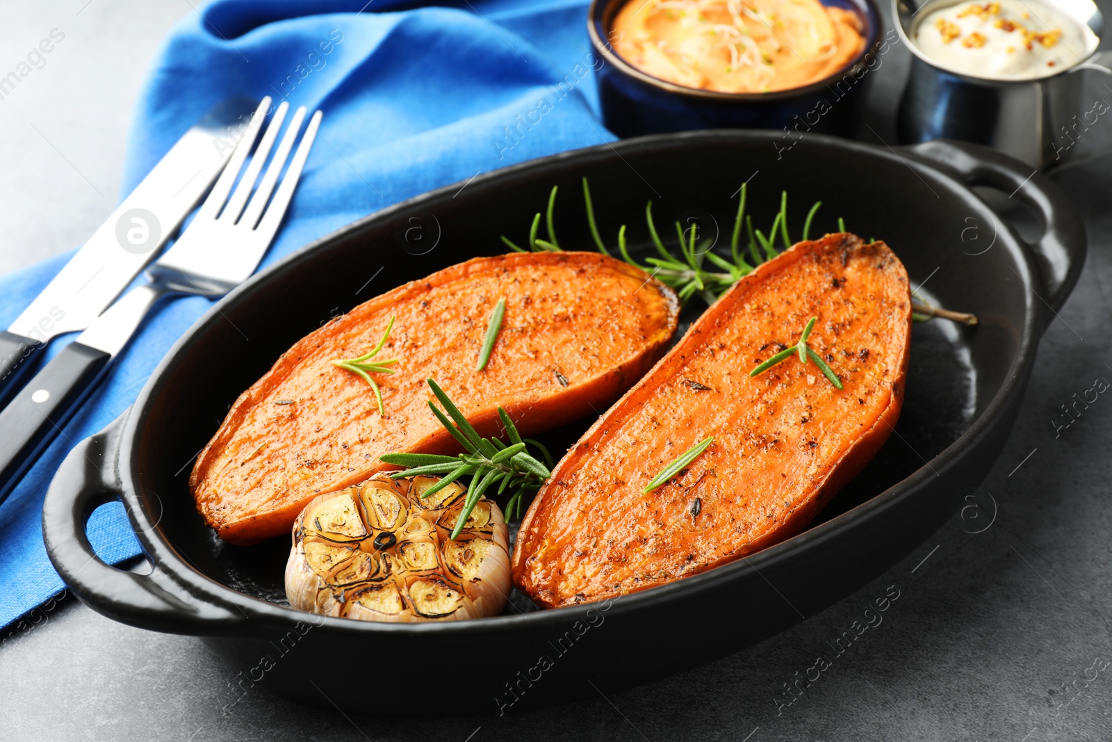 Photo of Halves of tasty cooked sweet potato with rosemary and garlic in baking dish served on grey table, closeup