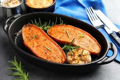 Photo of Halves of tasty cooked sweet potato with rosemary and garlic in baking dish served on grey table, closeup