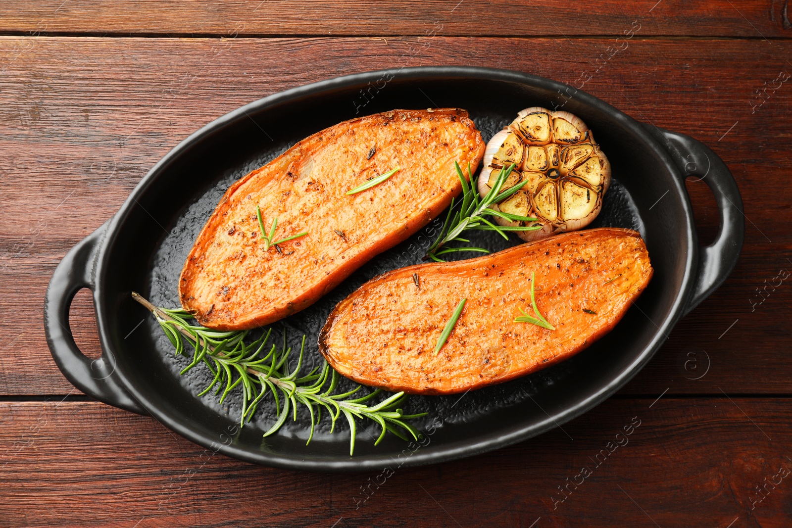 Photo of Halves of tasty cooked sweet potato with rosemary and garlic in baking dish on wooden table, top view