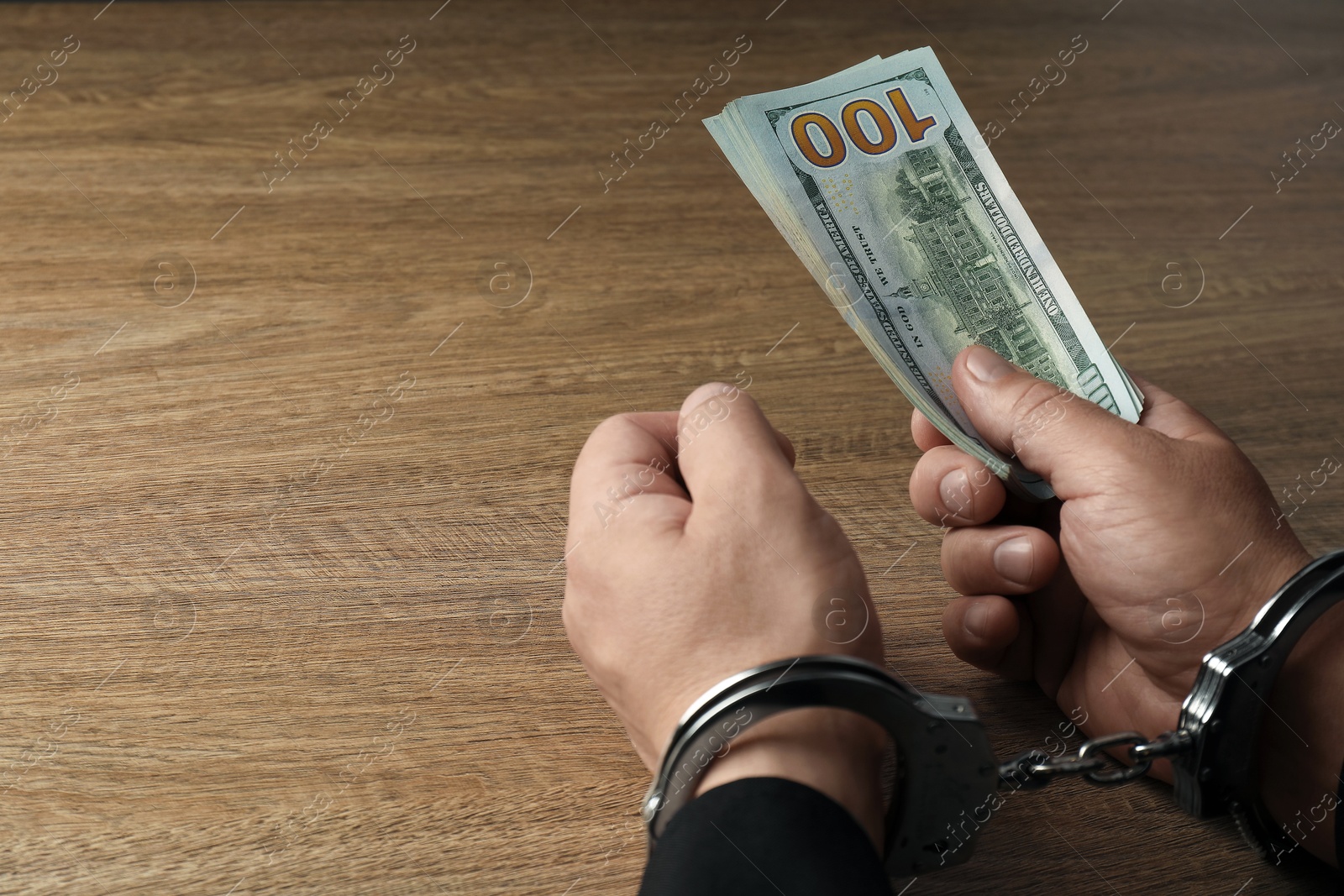 Photo of Corruption. Man in handcuffs with dollar banknotes at wooden table, closeup. Space for text