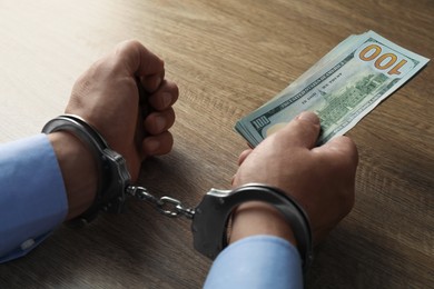 Photo of Corruption. Man in handcuffs holding dollar banknotes at wooden table, closeup