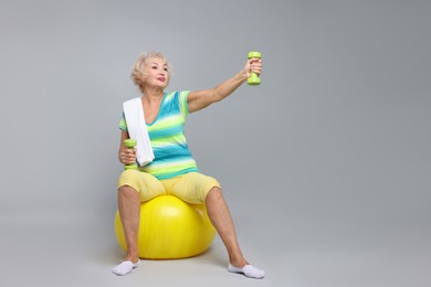 Photo of Senior woman exercising with fitness ball, dumbbells and towel on light grey background, space for text