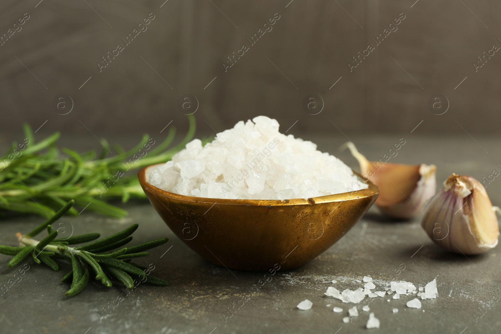 Photo of Sea salt in bowl, rosemary and garlic on grey table, closeup