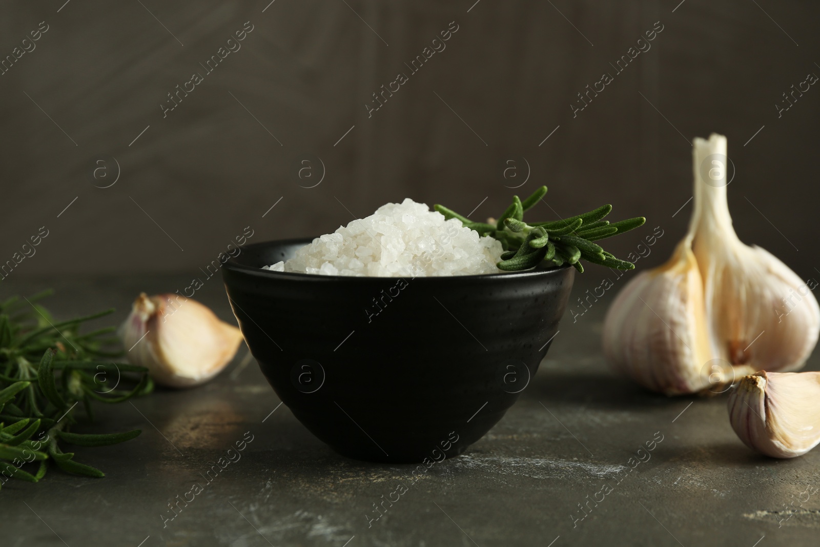 Photo of Sea salt in bowl, rosemary and garlic on grey table, closeup