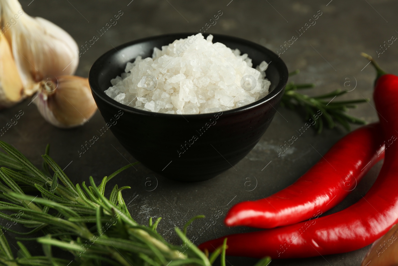 Photo of Sea salt in bowl, rosemary, chili peppers and garlic on grey table, closeup