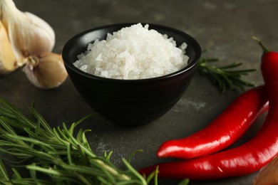 Photo of Sea salt in bowl, rosemary, chili peppers and garlic on grey table, closeup