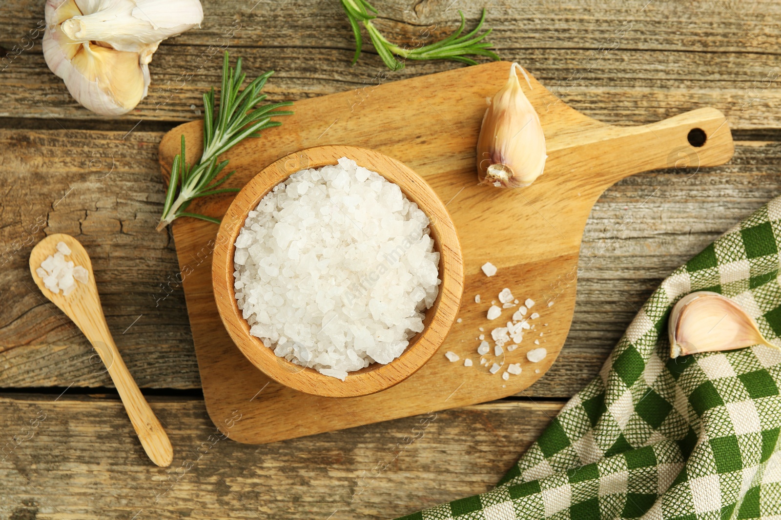 Photo of Sea salt in bowl, rosemary and garlic on wooden table, top view