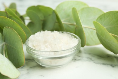 Photo of Sea salt in glass bowl and eucalyptus leaves on white marble table, closeup