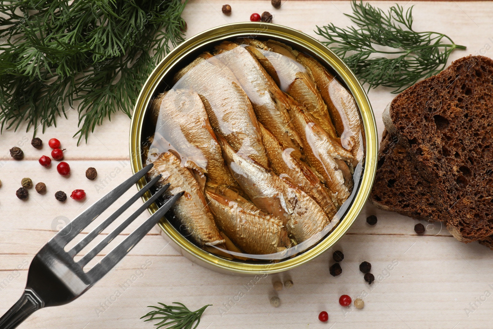 Photo of Tasty sprats in tin can, dill, bread and peppercorns on wooden table, top view