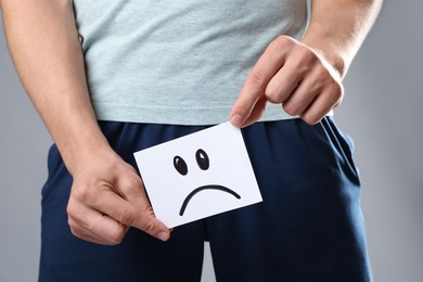 Photo of STD (sexually transmitted diseases). Man holding sticky note with drawn sad face on grey background, closeup