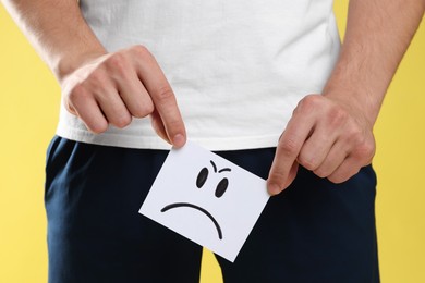 Photo of STD (sexually transmitted diseases). Man holding sticky note with drawn angry face on yellow background, closeup