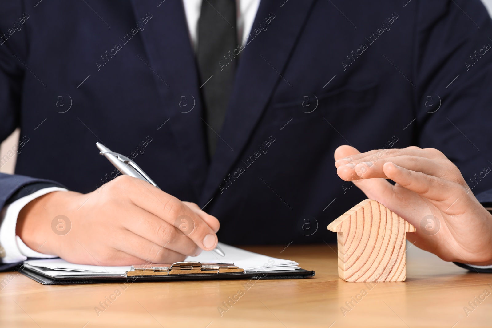 Photo of Property insurance. Real estate agent protecting house figure while working at wooden table, closeup