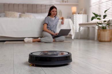 Photo of Young woman using laptop at home, focus on robotic vacuum cleaner