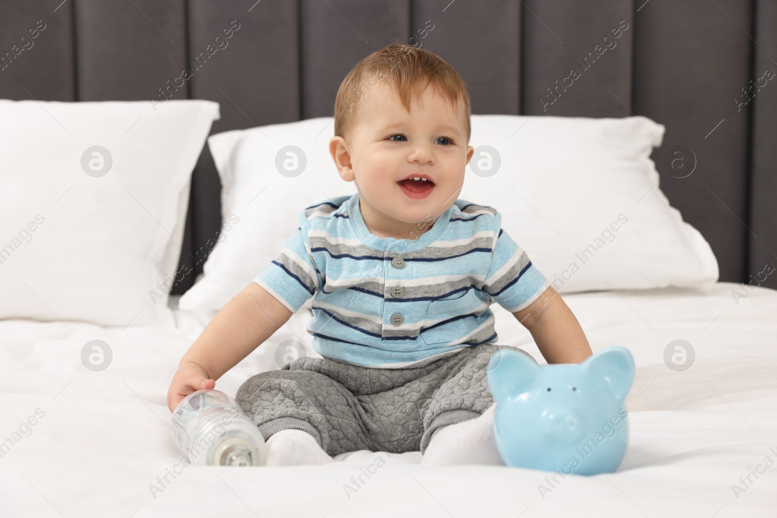 Photo of Cute little baby with money in bottle and piggybank on bed at home