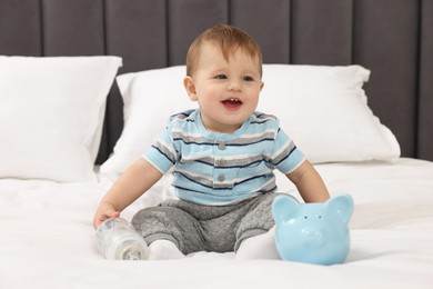 Photo of Cute little baby with money in bottle and piggybank on bed at home