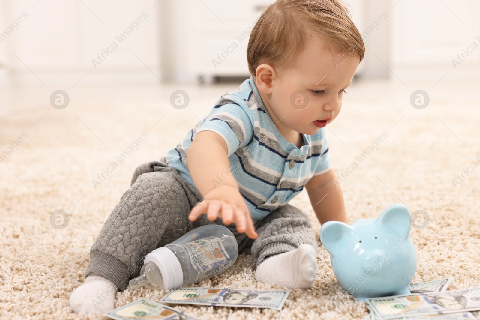 Photo of Little baby with bottle, money and piggybank on floor at home