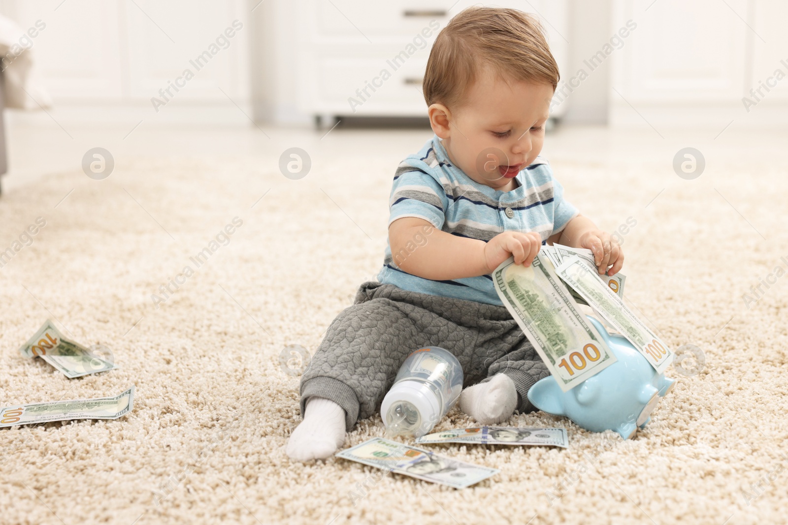 Photo of Little baby with bottle, money and piggybank on floor at home