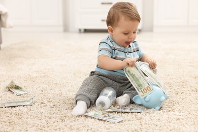 Photo of Little baby with bottle, money and piggybank on floor at home