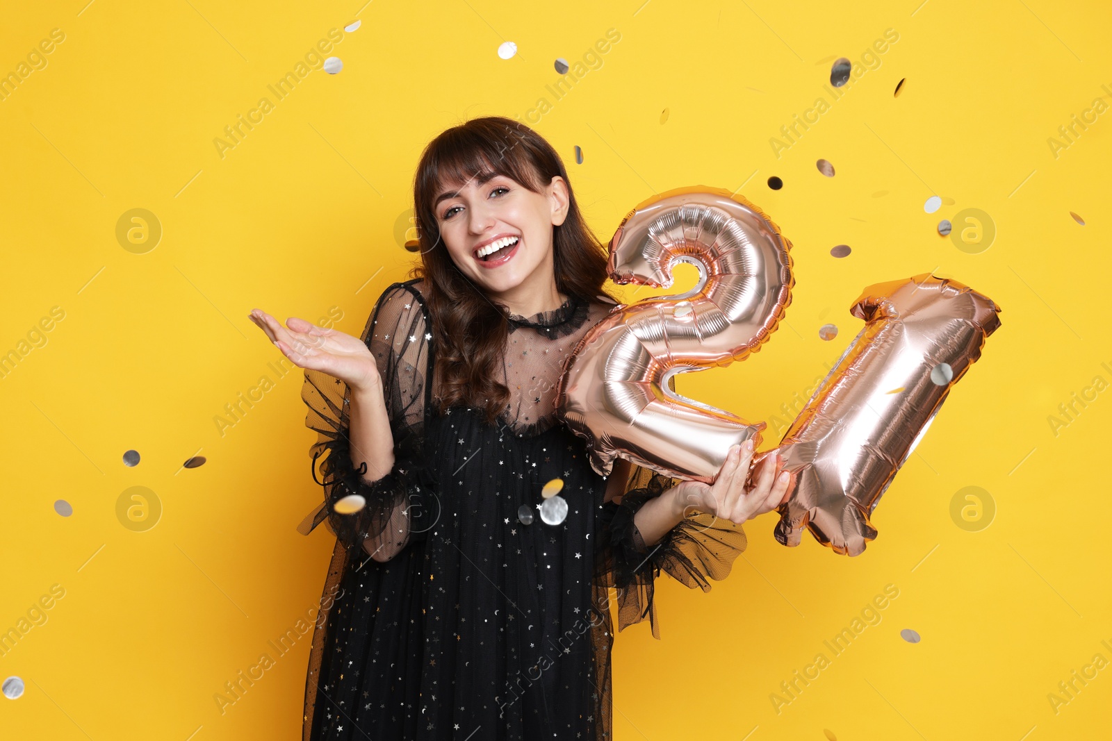 Photo of Coming of age party - 21st birthday. Young woman holding number shaped balloons on yellow background with confetti