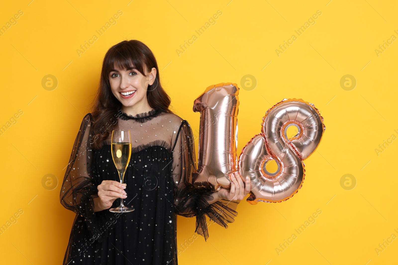 Photo of Coming of age party - 18th birthday. Young woman holding number shaped balloons and glass of wine on yellow background