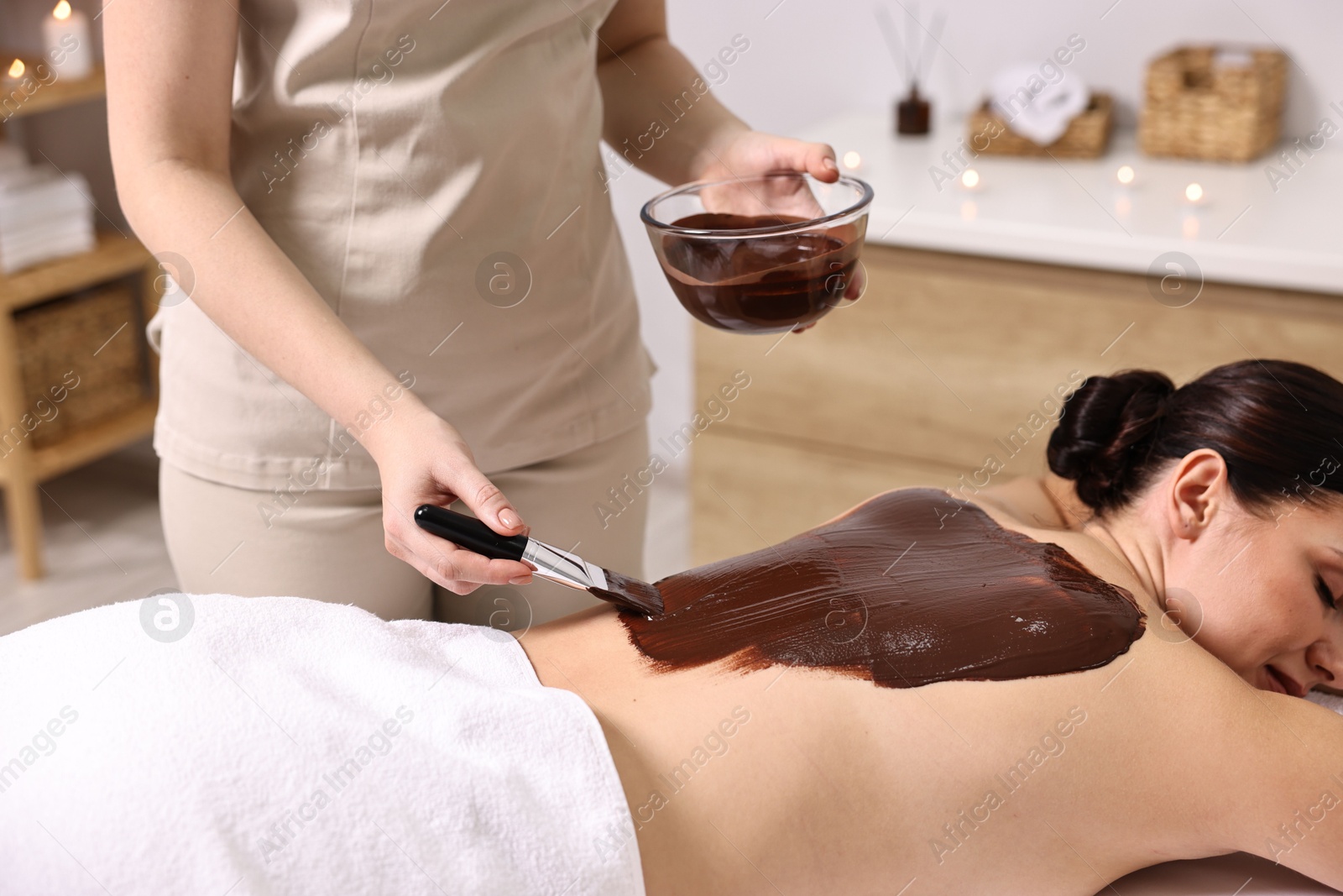 Photo of Chocolate body wrap. Spa worker applying mask onto woman's back in salon, closeup