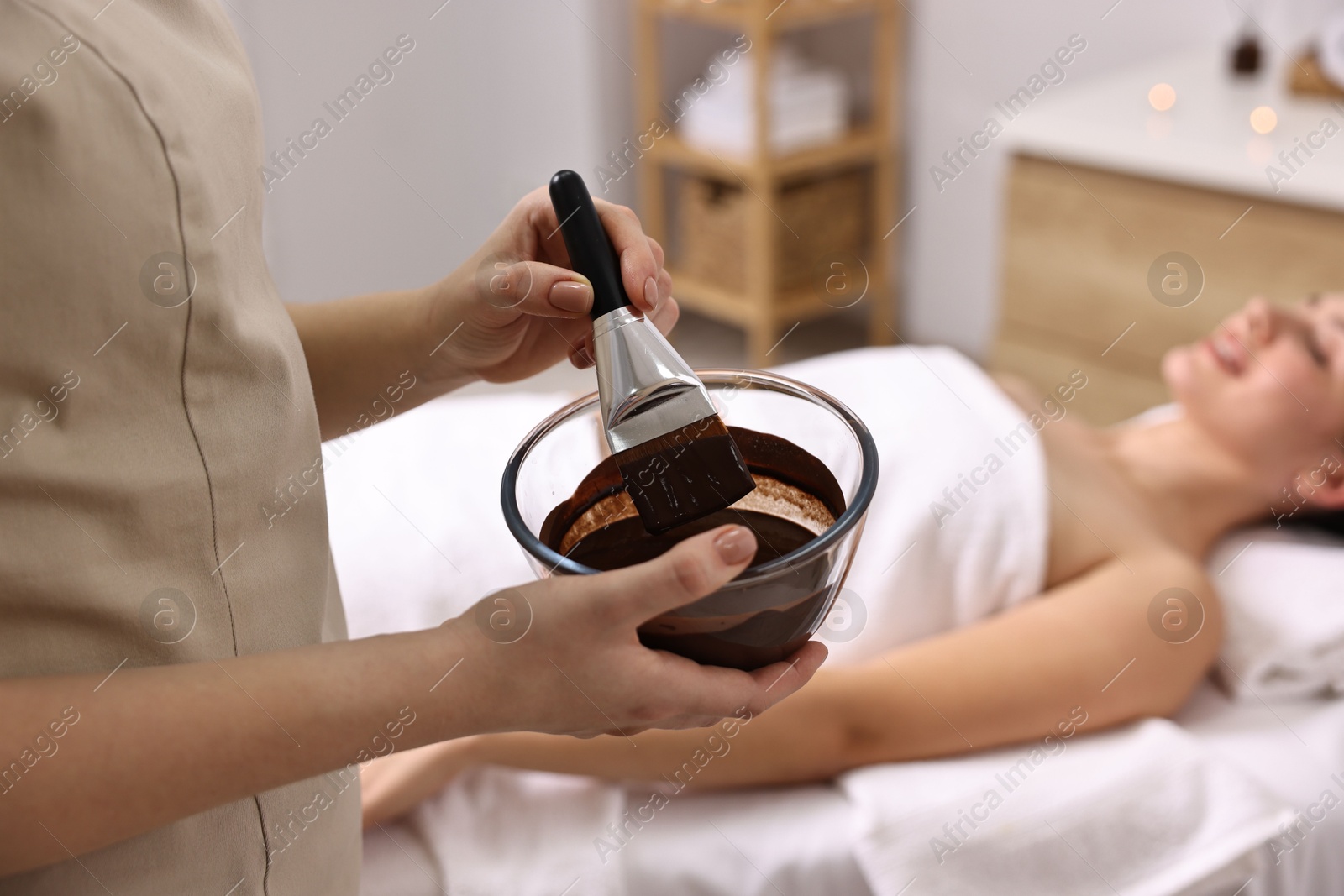 Photo of Spa worker with cosmetic product for chocolate body wrap near woman in salon, closeup