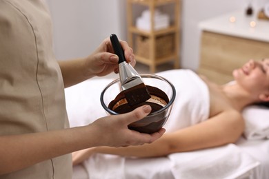 Photo of Spa worker with cosmetic product for chocolate body wrap near woman in salon, closeup